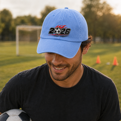 Man holding a football on a practice pitch wearing a light blue washed baseball cap with an embroidered flaming 2026 soccer ball design