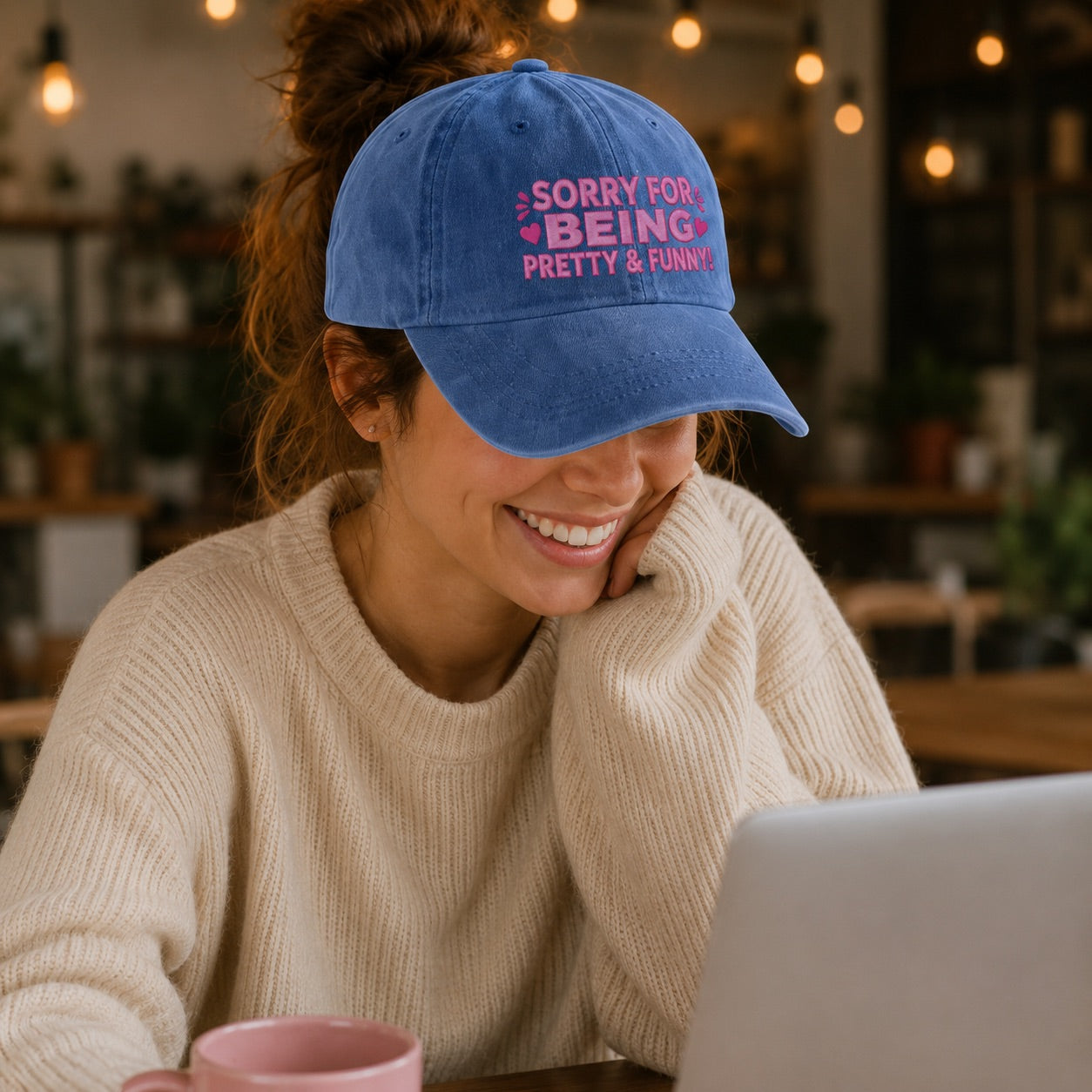 Woman wearing a blue cap with text, smiling and using a laptop in a cozy indoor setting.