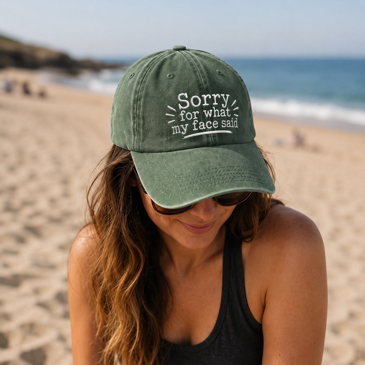 Woman on a beach wearing a green cap with text, sitting by the ocean.