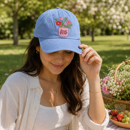 Woman wearing a blue cap with floral design and text, standing outdoors in a garden.
