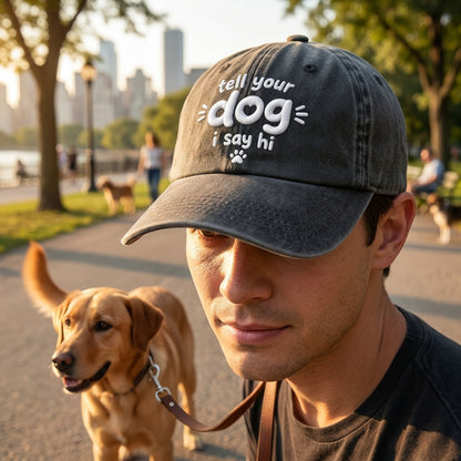 Man wearing a cap with 'tell your dog i say hi' text, standing with a dog in a park setting.