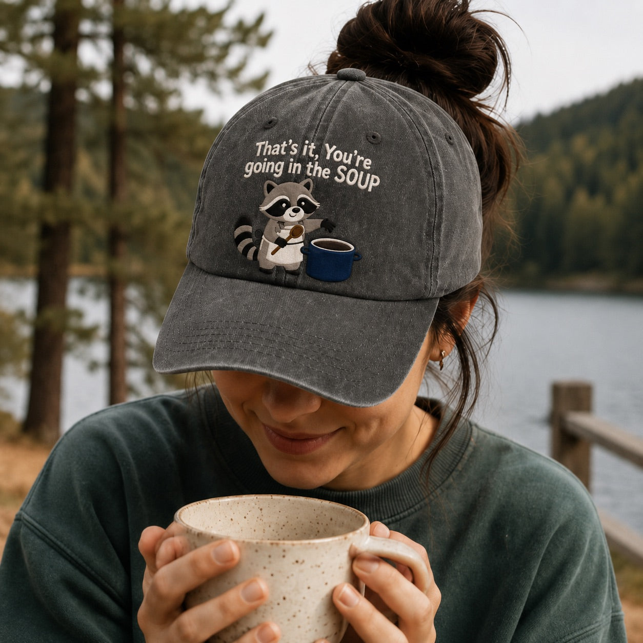 Person wearing a cap with a raccoon graphic and text, holding a mug outdoors by a lake.