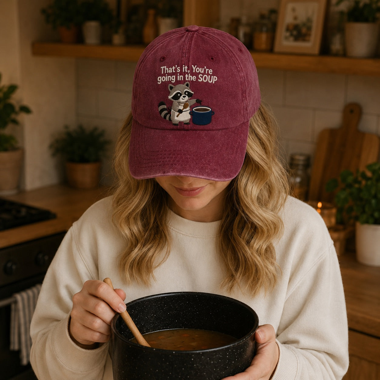 Person wearing a maroon cap with a raccoon graphic and text, holding a bowl of soup in a kitchen.