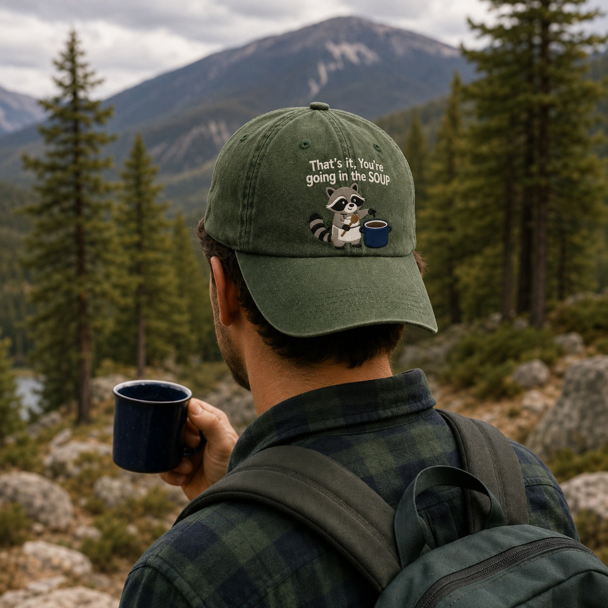 Person wearing a cap with a raccoon design and text, holding a mug in a mountainous forest setting.