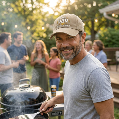 Man wearing a cap with 'This Dad Needs a Beer' text, standing by a grill with people in the background.