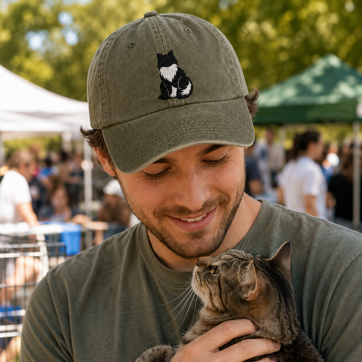 Man wearing an olive green washed cap with a detailed Tuxedo cat embroidery while holding a kitten