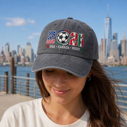 Woman with city skyline background wearing a grey washed baseball cap with embroidered 2026 tri-nation soccer tournament design