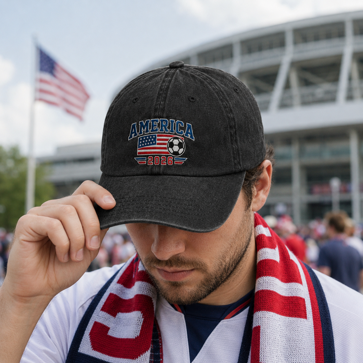 Man wearing a patriotic scarf outside a stadium and adjusting a black washed baseball cap with an embroidered America 2026 soccer and US flag design