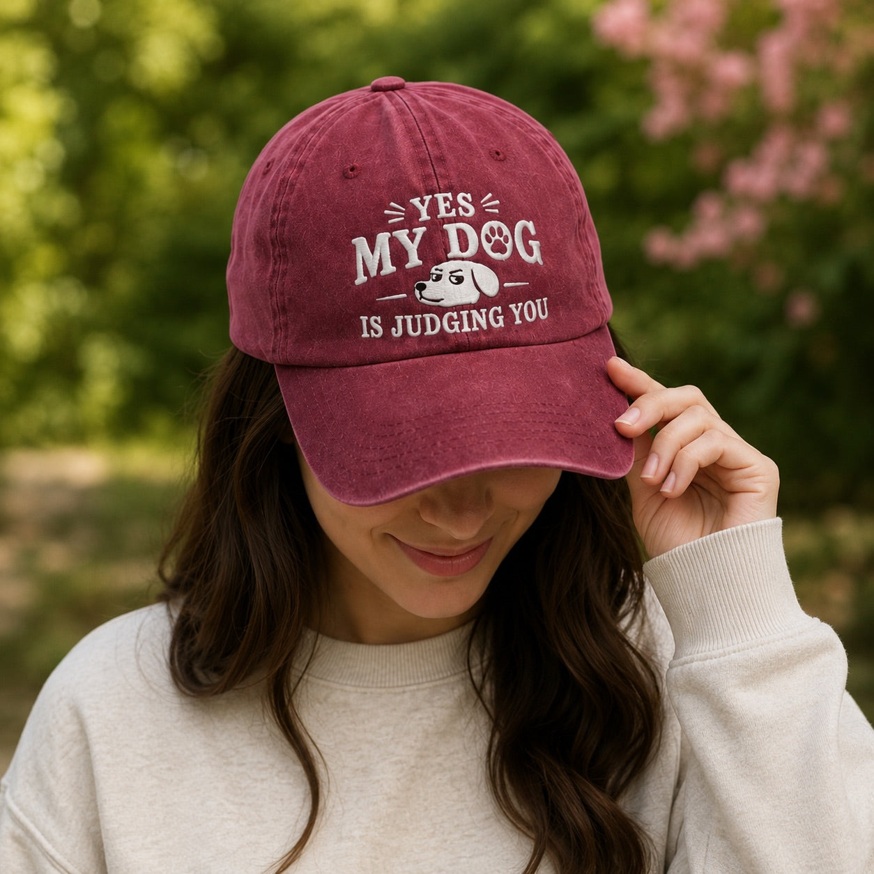 Person wearing a maroon cap with 'YES MY DOG IS JUDGING YOU' text and dog graphic, outdoors.